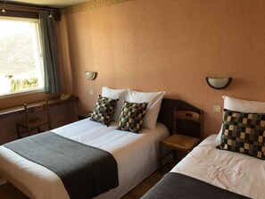A hotel room with two beds, a window with curtains, wall-mounted lamps, and a small table.