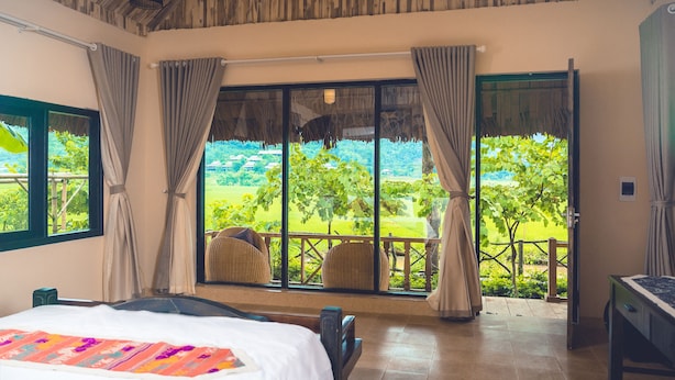 A room with a bed, a view of a lush green landscape, and a balcony with wicker furniture. - Mai Chau Sunrise Village (Mai Chau)