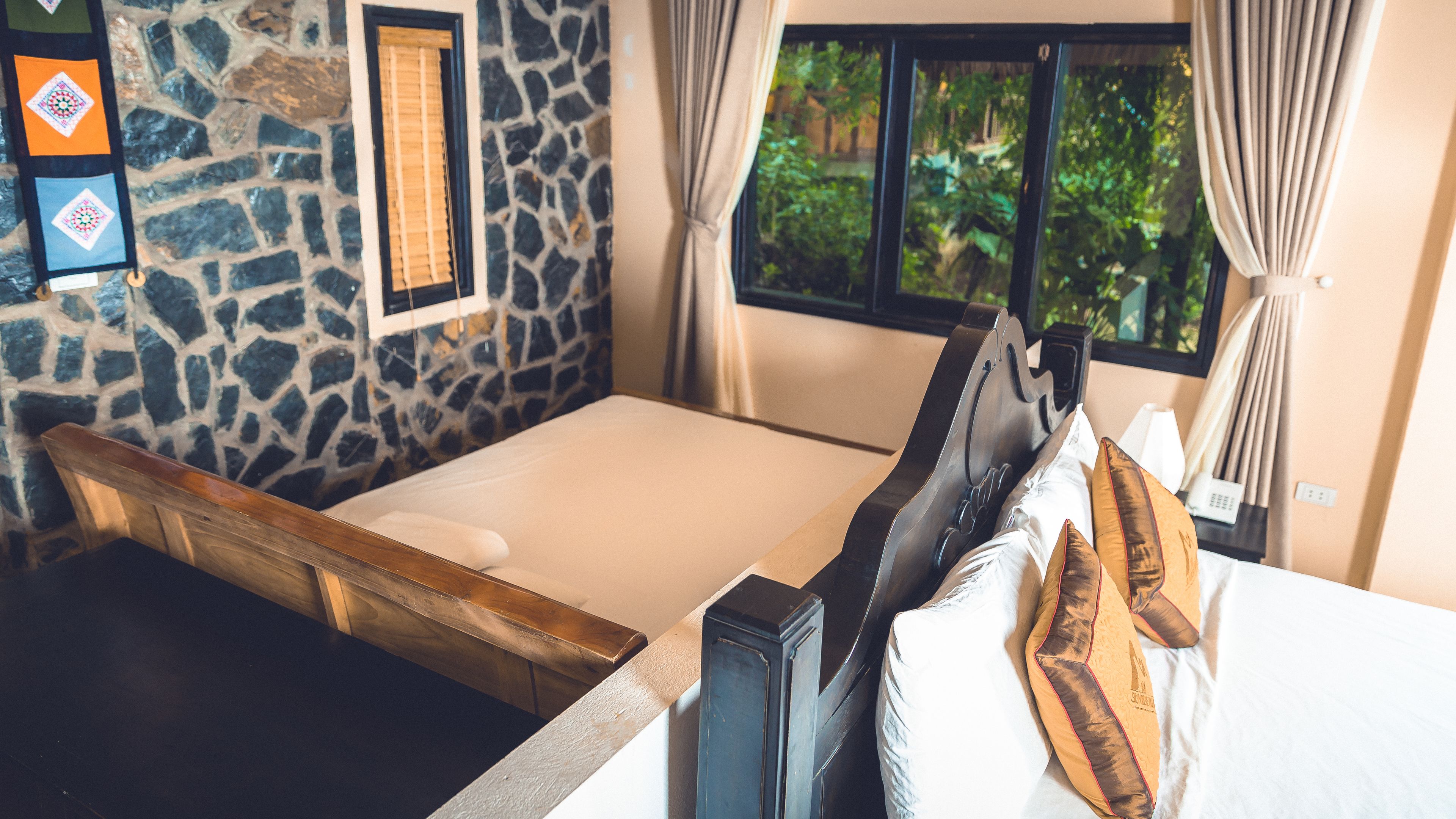 A room with a stone wall, a bed with a wooden headboard, a window with curtains, and a view of greenery outside.
