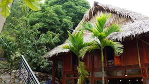 A wooden building with a thatched roof, surrounded by lush greenery and a stone staircase. - Mai Chau Sunrise Village (Mai Chau)