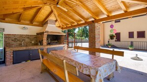 Ein Outdoor-Kochbereich mit Backofen aus Ziegelsteinen, einer Holzbänkl und einem Tisch unter einer Holzdachkonstruktion.