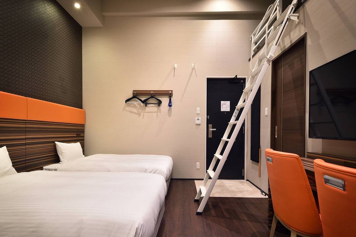 Une chambre d’hôtel avec deux lits, une télévision, une échelle et des chaises oranges.