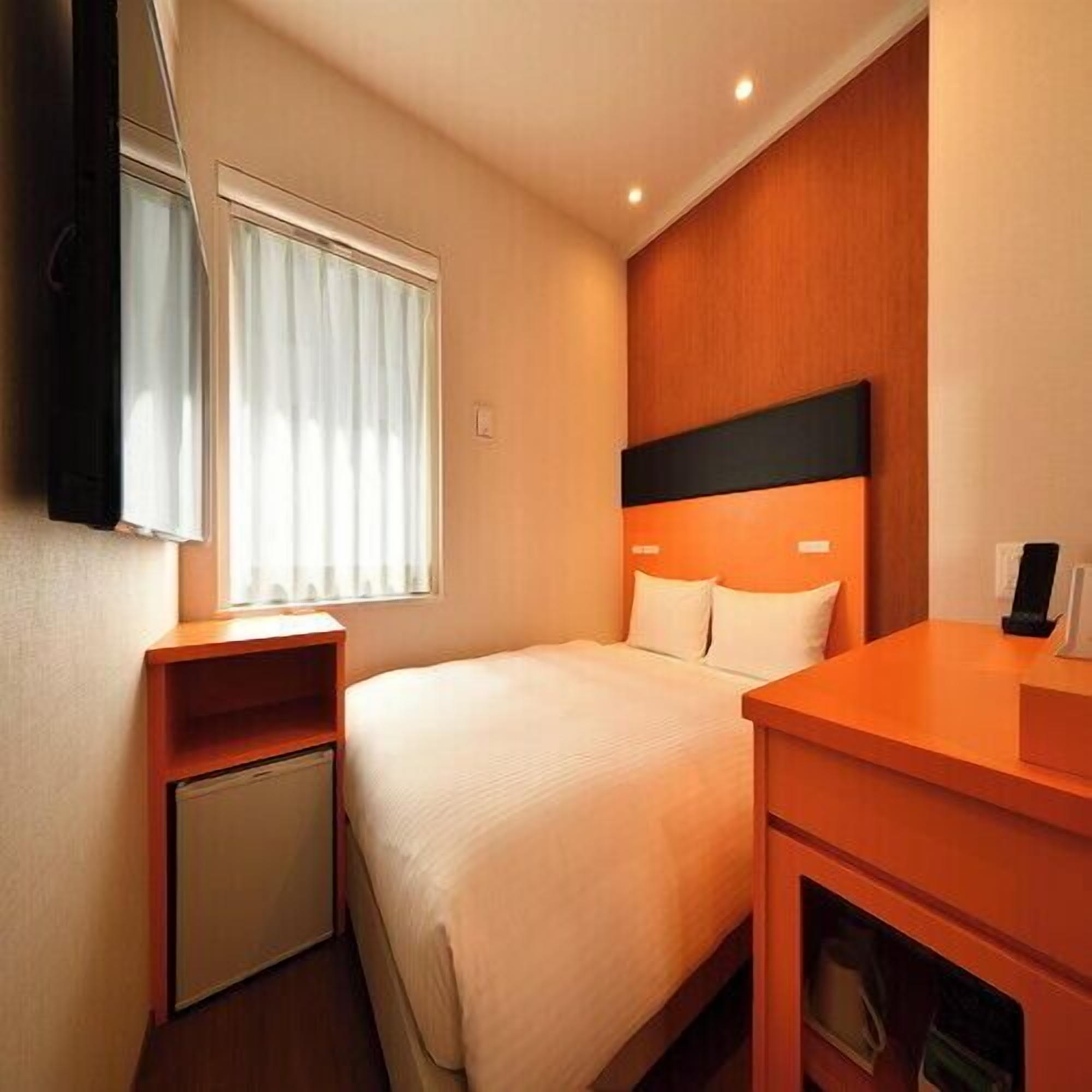 A hotel room with a bed, a bedside table, a small cabinet, a window with blinds, and a wall-mounted TV.