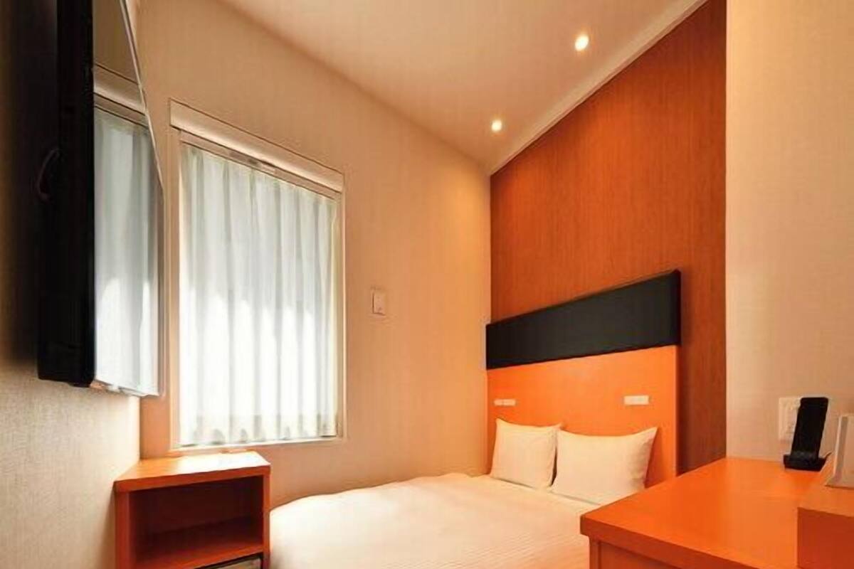 Une chambre d’hôtel comprenant un lit, une table de chevet, une petite armoire, une fenêtre avec des stores et un téléviseur fixé au mur.