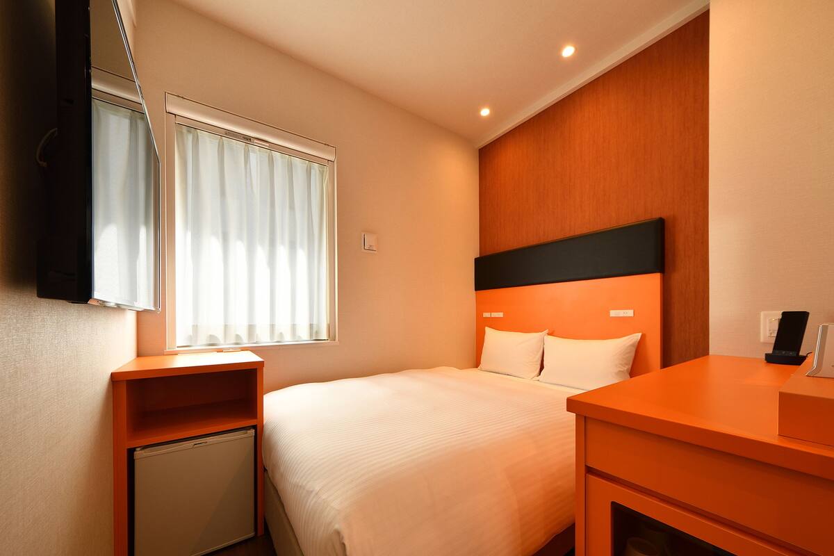 Une chambre d’hôtel avec un lit, un petit bureau, un téléviseur fixé au mur et une fenêtre avec des rideaux.