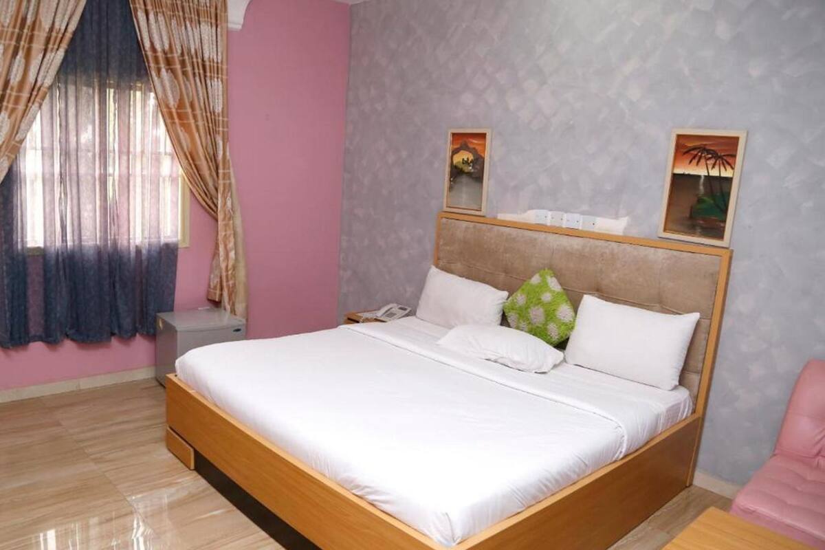 A neatly made bed in a room with a pink wall, a patterned headboard, and a window with sheer curtains.