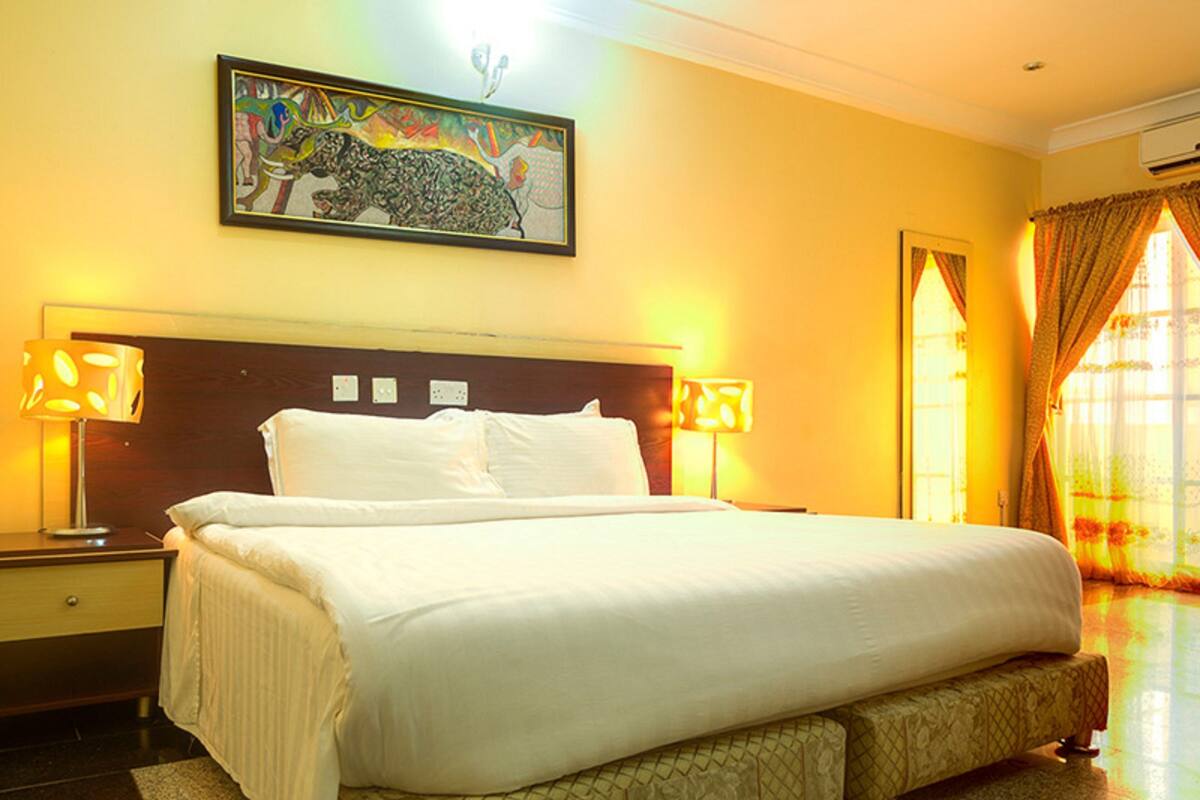 A hotel room with a large bed, two bedside lamps, a nightstand, a framed artwork above the bed, and a window with curtains.