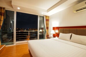 A hotel room with a large bed, a nightstand with a lamp, a window with curtains, and an air conditioning unit. - Jomtien Hisotel (Pattaya)