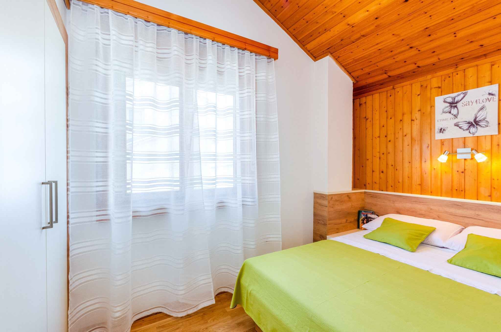 Ein Schlafzimmer mit einem Bett, grünen Kissen, einem Holz-Kopfteil, einer Wandleuchte und einem Fenster mit transparenten Vorhängen.