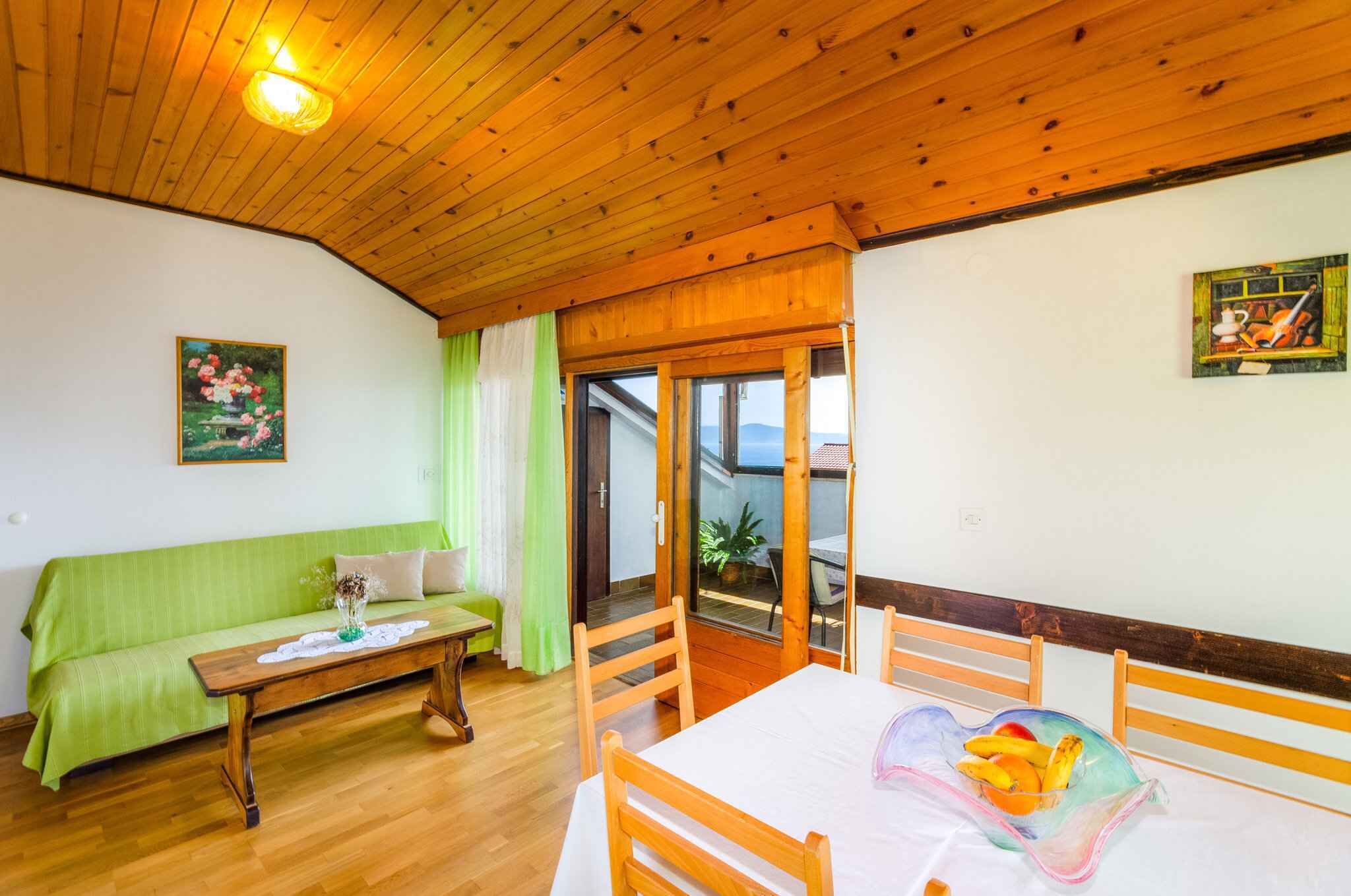 Una habitación acogedora con techo de madera, un sofá verde, una mesa de madera con un jarrón y una mesa de comedor con fruta.