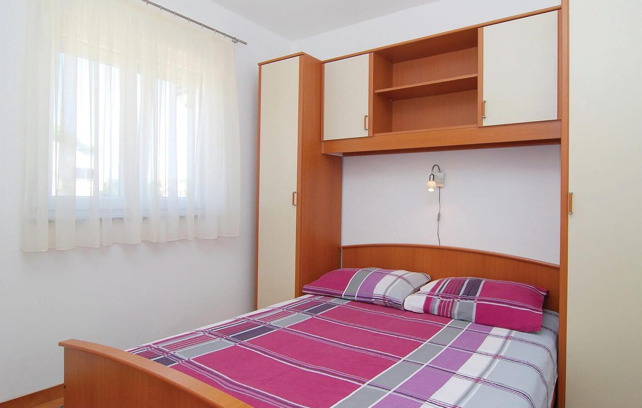 A bedroom with a bed, a window with sheer curtains, a bedside lamp, and built-in shelving above the bed.