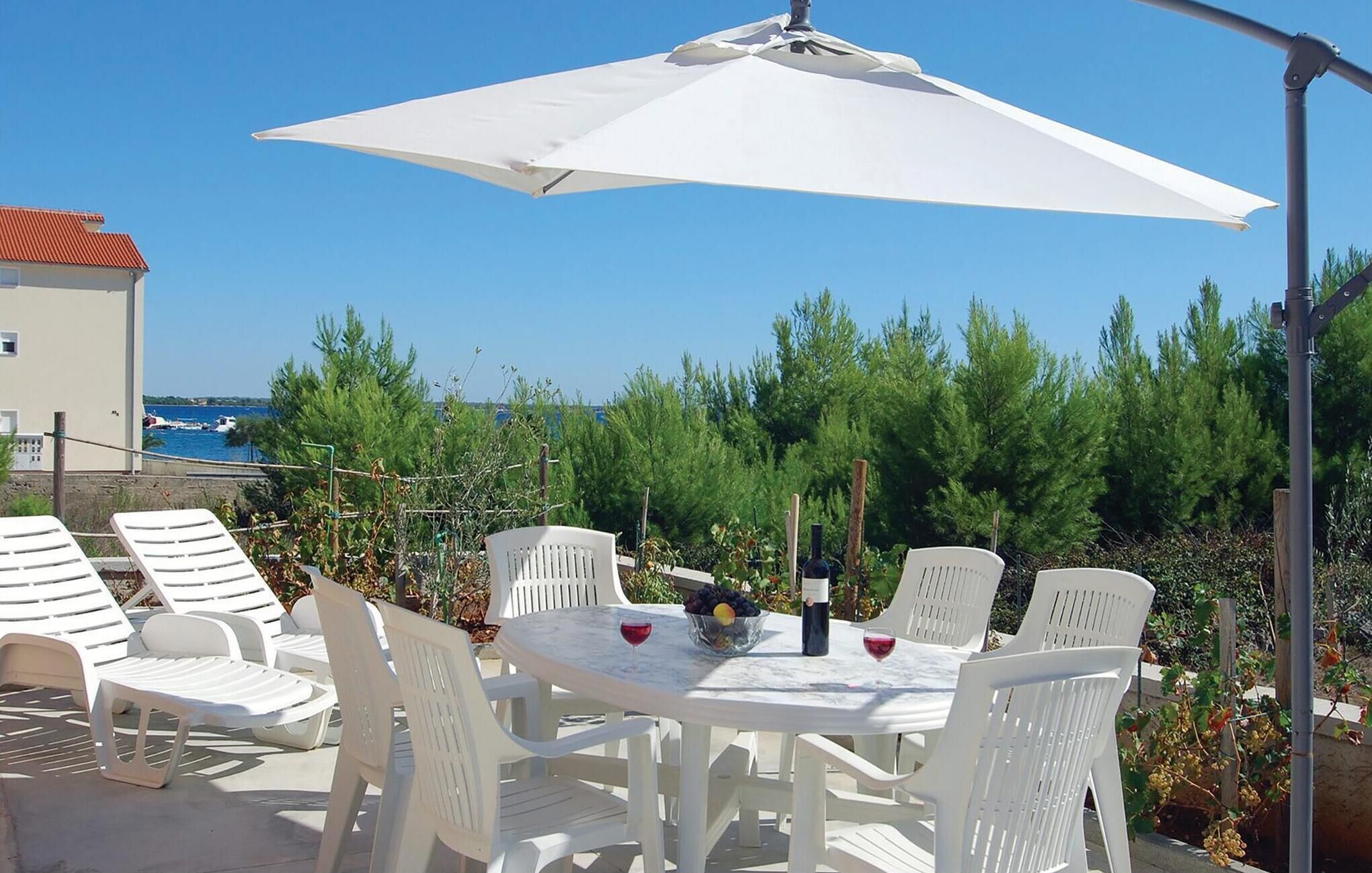 Een terras met witte plastic stoelen en een tafel, een witte parasol, en uitzicht op zee en bomen in de achtergrond.