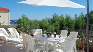 Un espace extérieur aménagé avec des chaises et une table en plastique blanc, un parasol blanc, et une vue sur la mer et les arbres en arrière-plan.