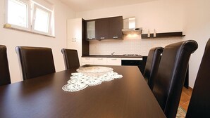 Un coin repas avec une table en bois et quatre chaises, une cuisine à l’arrière-plan et une fenêtre avec des stores.