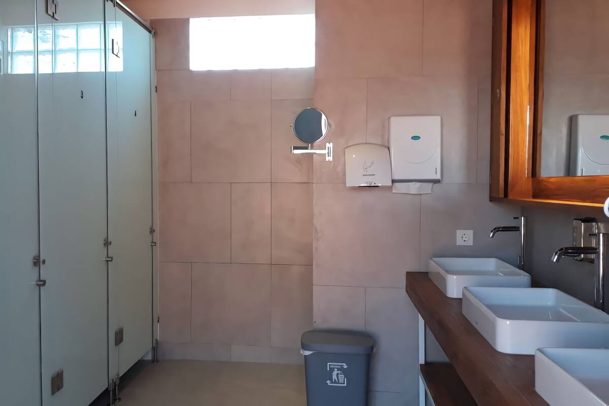 A bathroom with a row of lockers, a hand dryer, a trash bin, and a sink with a mirror above it.