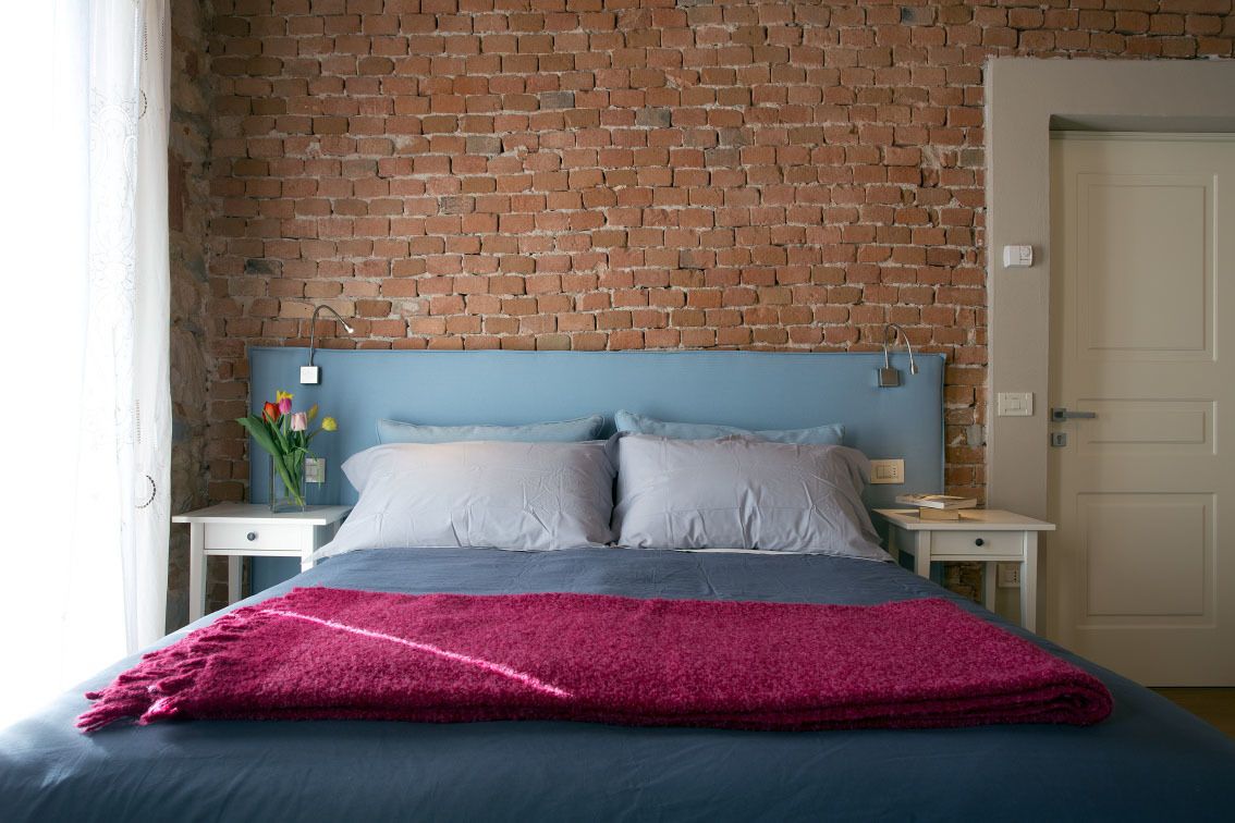 Une chambre avec un mur en briques, un lit avec une literie bleue et grise, un plaid rose et une table de chevet ornée d’un vase de fleurs.