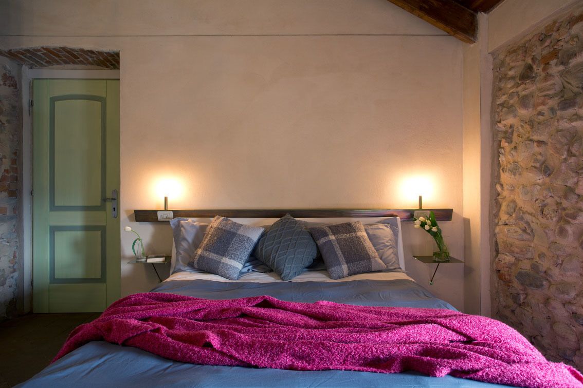 A bedroom with a bed, pillows, a blanket, bedside lamps, a nightstand, and a stone wall.