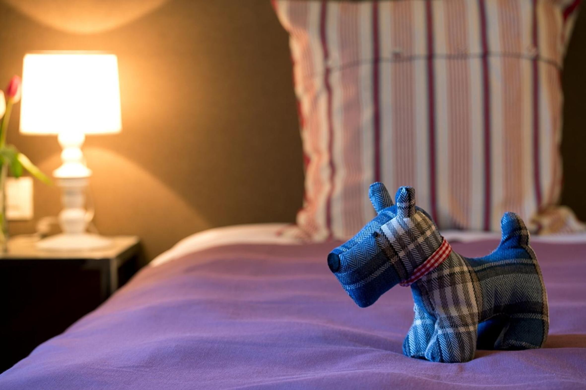 En seng med et lilla sengetæppe og en lille hundefigur ovenpå.