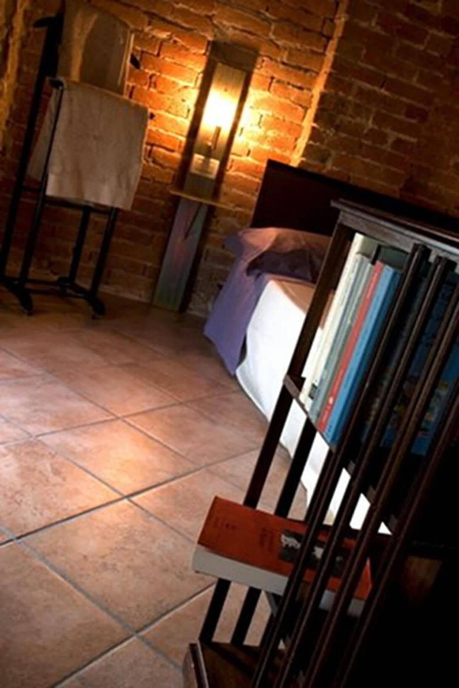 Una habitación con pared de ladrillo, una cama con cabecera blanca, una mesita de noche y una estantería llena de libros.