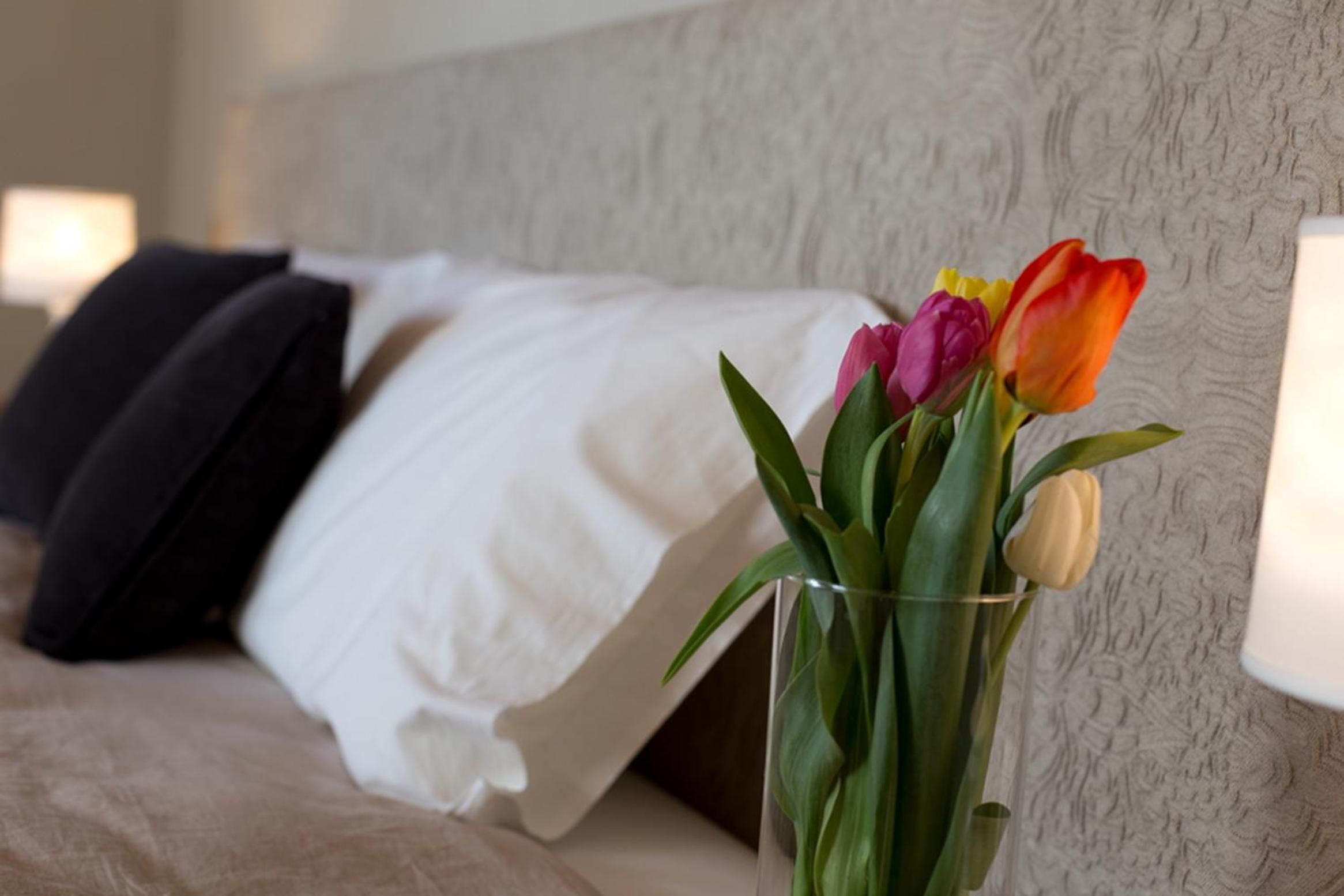 A bed with white and black pillows, a glass vase with tulips, and a textured wall in the background.
