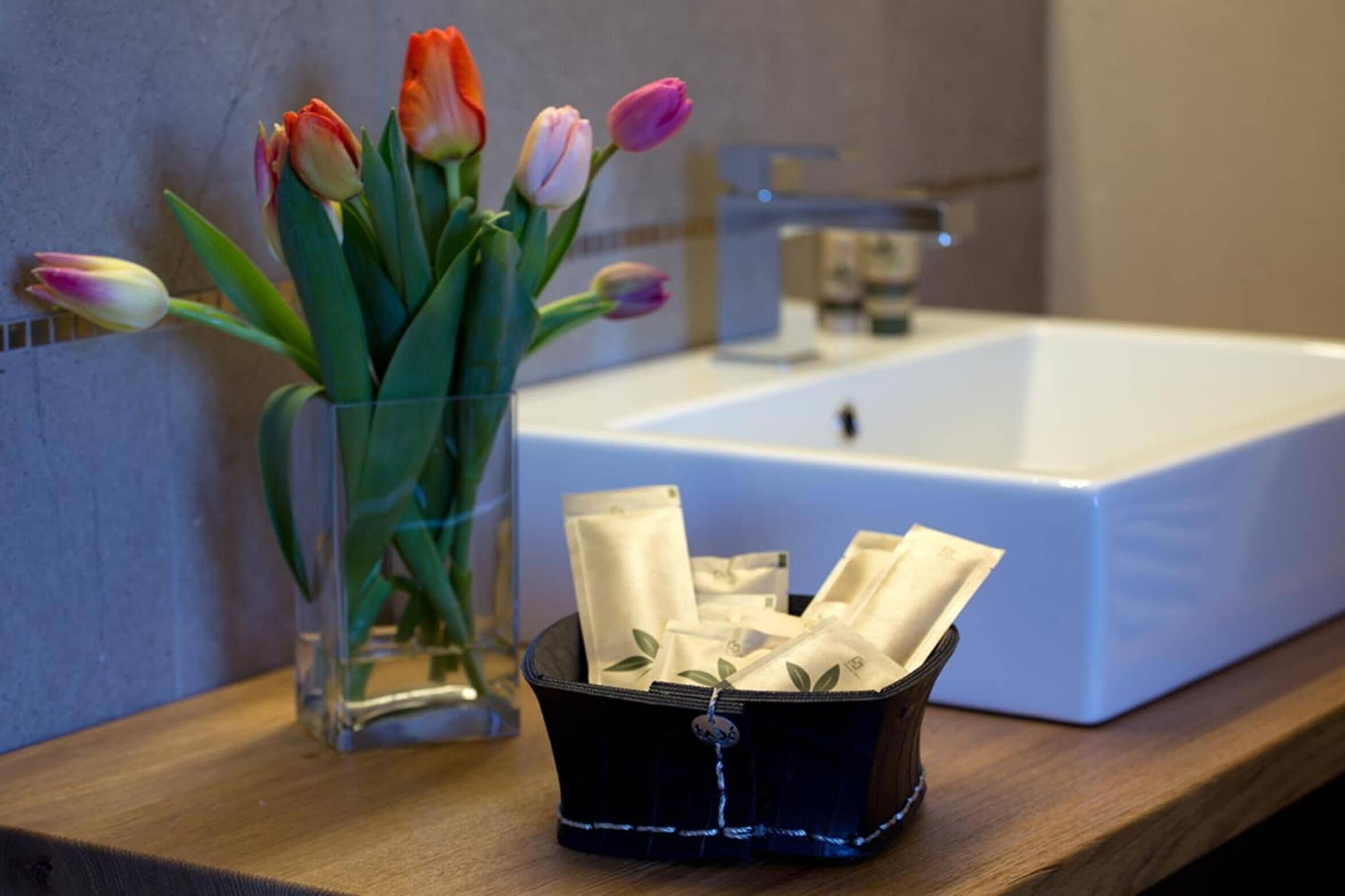 Un bagno con un lavandino bianco, un vaso di vetro con tulipani e un contenitore nero con fazzeletti.