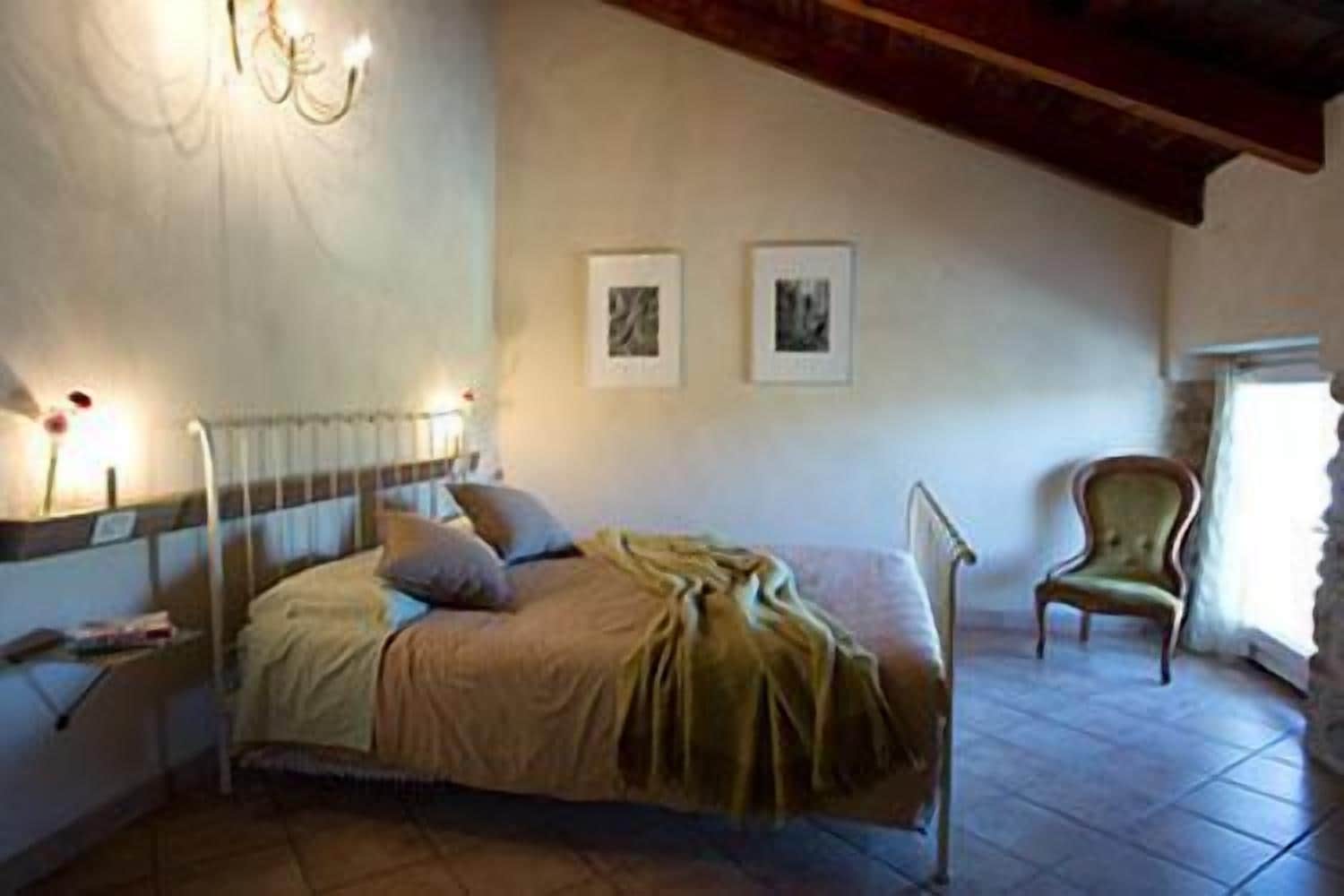 Un dormitorio con cama, mesita de noche, lámpara, silla y dos cuadros en la pared.