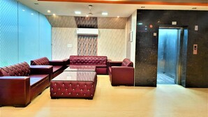 Un hall d’entrée avec un espace salon comprenant des canapés rouges recouverts de tissu et une table basse en verre. Un climatiseur est fixé au mur, et un ascenseur est visible sur la droite.