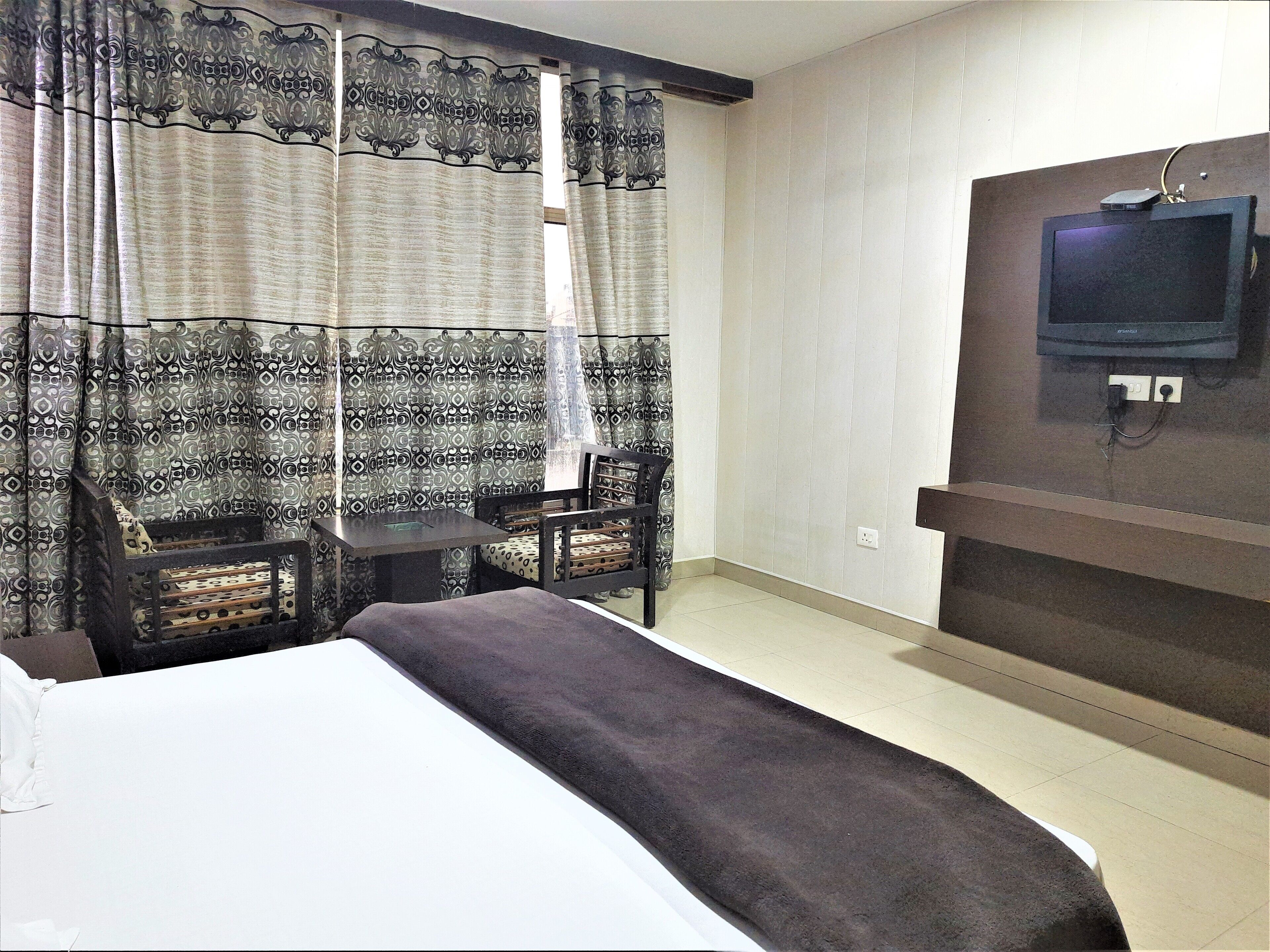Ein Hotelzimmer mit einem Bett, einem Schreibtisch, zwei Stühlen, einem Fernseher und gemusterten Vorhängen.