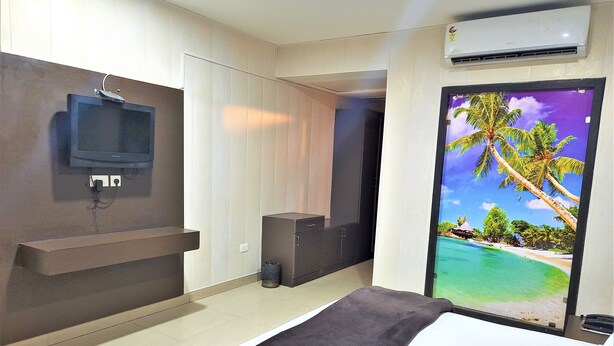 Une chambre d’hôtel moderne, équipée d’un lit, d’un téléviseur fixé au mur, d’une petite armoire et d’une porte ornée d’une fresque aux motifs tropicaux.