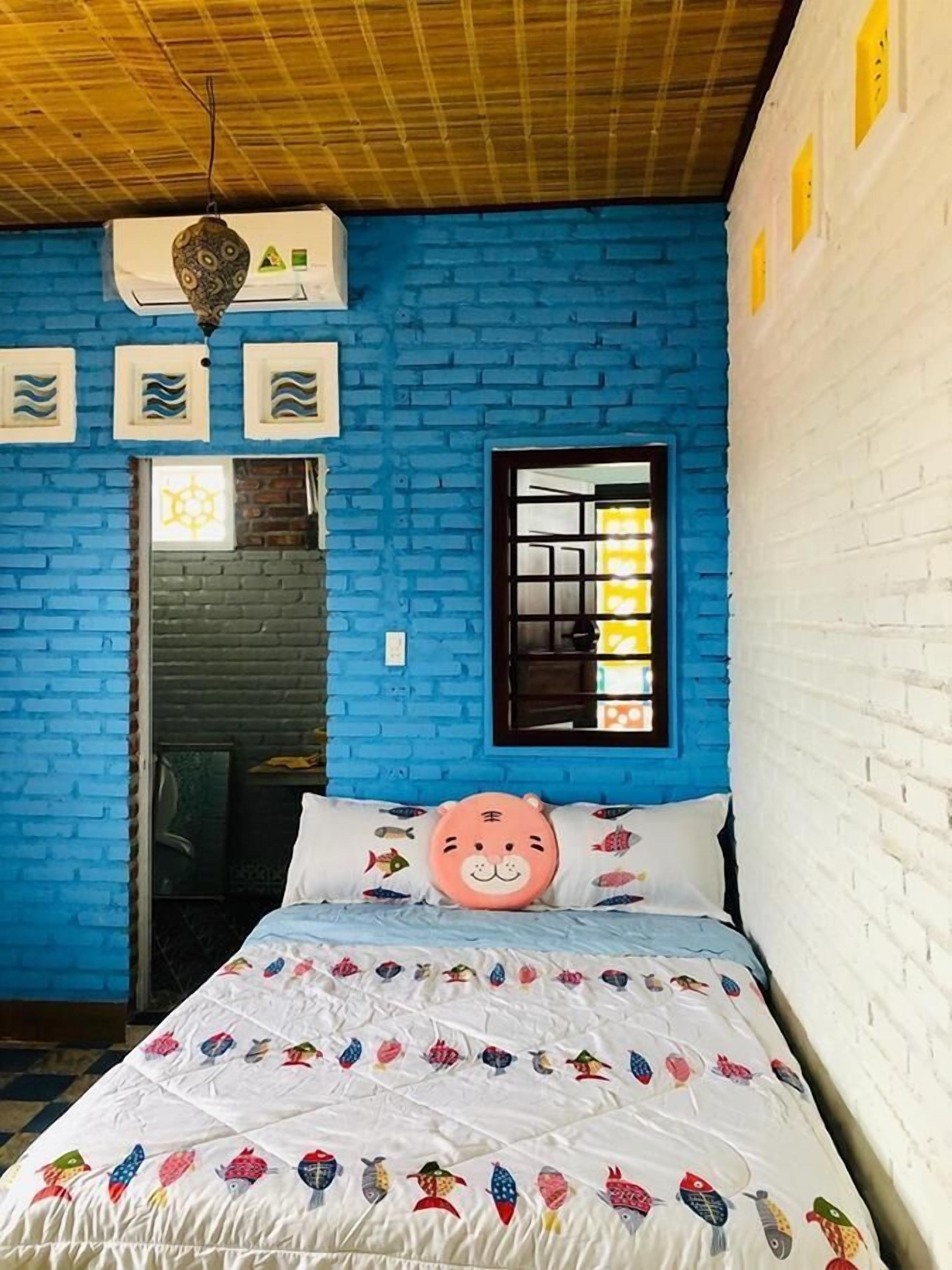 Une pièce aux murs en briques bleues, avec un plafond en bois, un lit recouvert d’une couette blanche ornée de motifs de poissons, un oreiller rose et une fenêtre à carreaux.