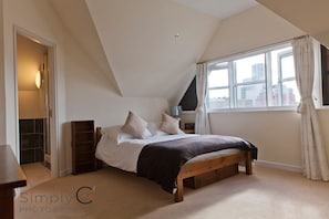 A bedroom with a sloped ceiling, a bed with a wooden frame, a bedside table, a lamp, and a window with curtains.