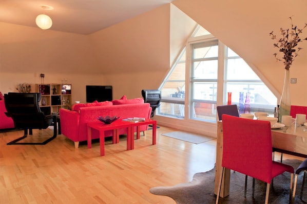 A modern living room with a red sofa, a black chair, a dining table with red chairs, and a large window offering a view of the city.