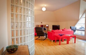 A modern living room with a red sofa, a black chair, and a wooden coffee table. A large window provides natural light, and a TV is mounted on the wall.