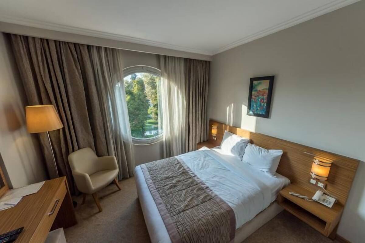 A hotel room with a bed, a desk, a chair, a nightstand, a lamp, a window with curtains, and a painting on the wall.