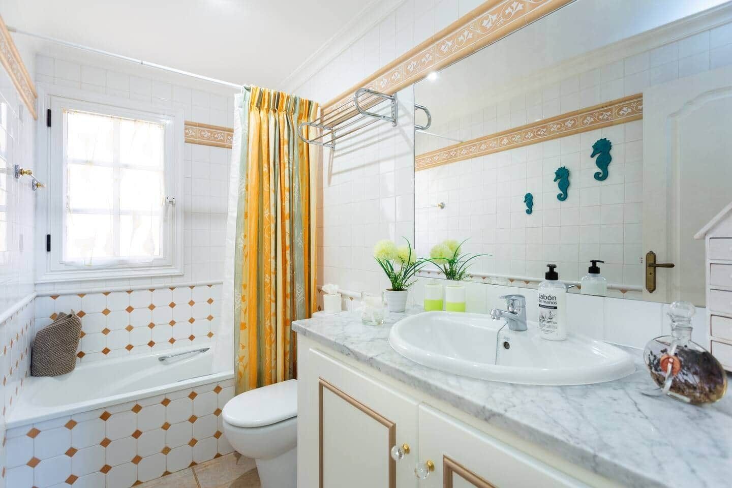 Un baño con bañera, inodoro y lavamanos con espejo. Las paredes están cubiertas de azulejos decorativos y hay una ventana con cortinas.