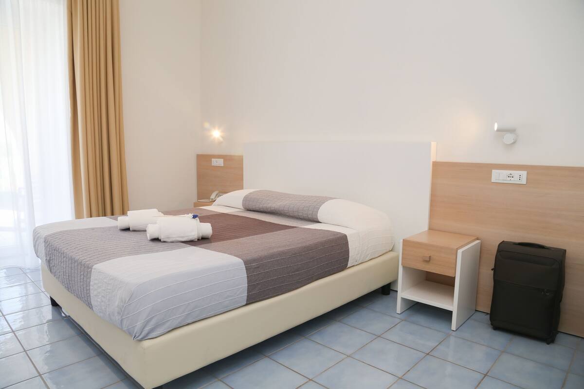 A neatly made bed with a brown and white striped blanket, a bedside table with a lamp, and a suitcase against the wall.