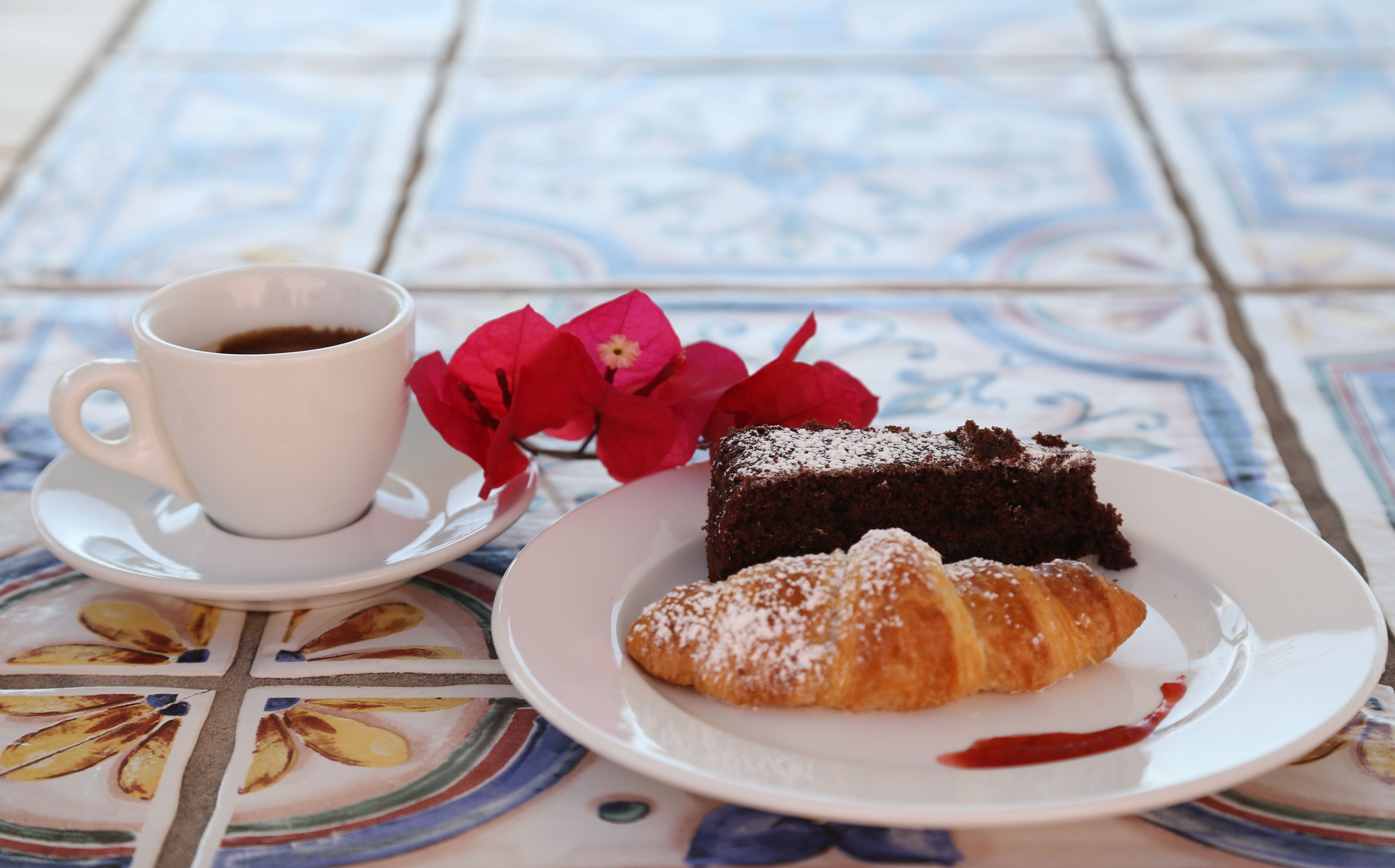 Uma mesa com uma xícara de café, uma fatia de bolo, um croissant e flores vermelhas.