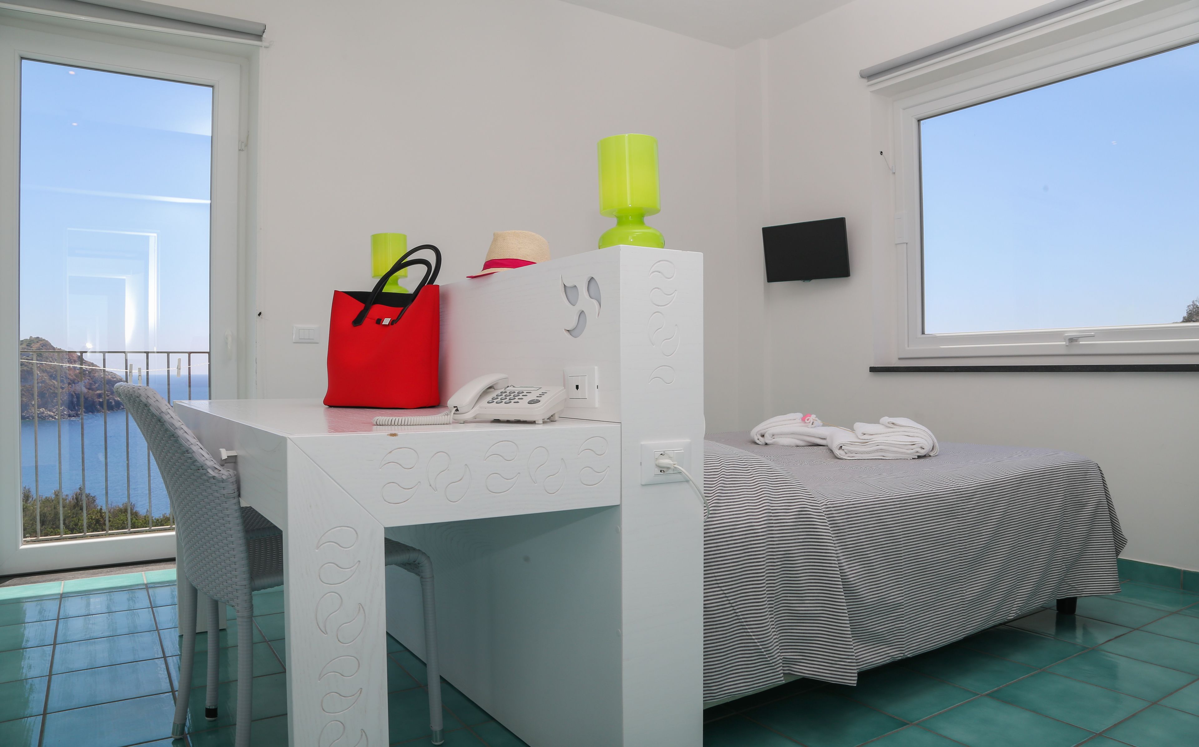 A room with a white desk, a red bag, a phone, and a bed with a striped bedspread.