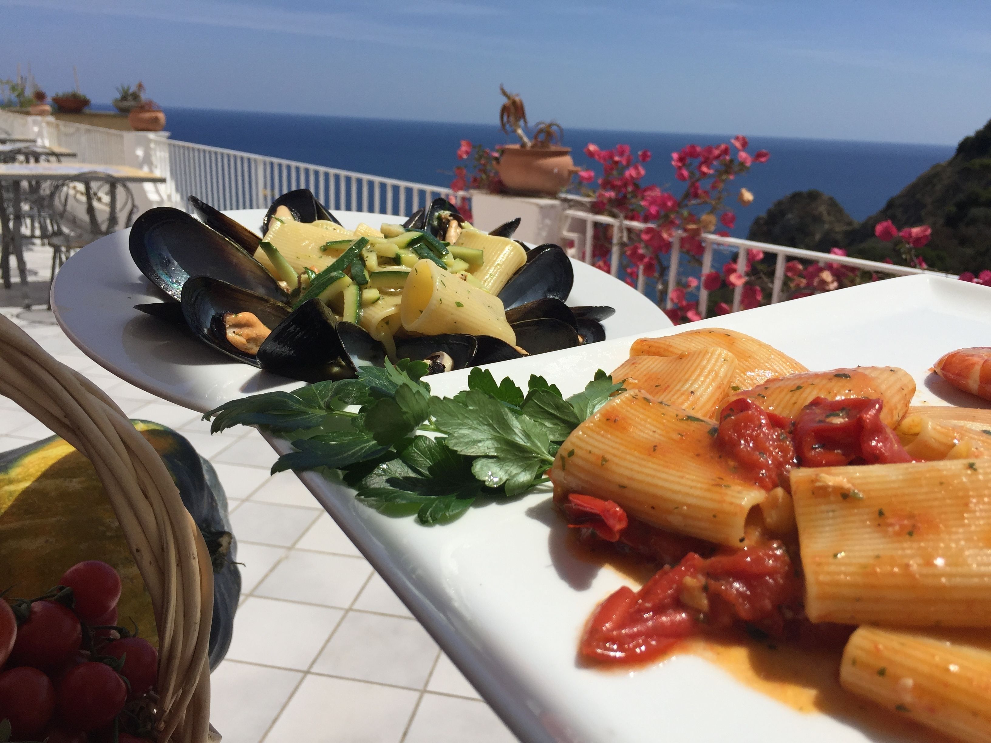A dining table with plates of pasta and mussels, set on a balcony with a view of the sea.