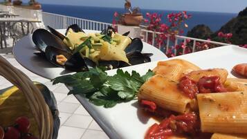 A dining table with plates of pasta and mussels, set on a balcony with a view of the sea.