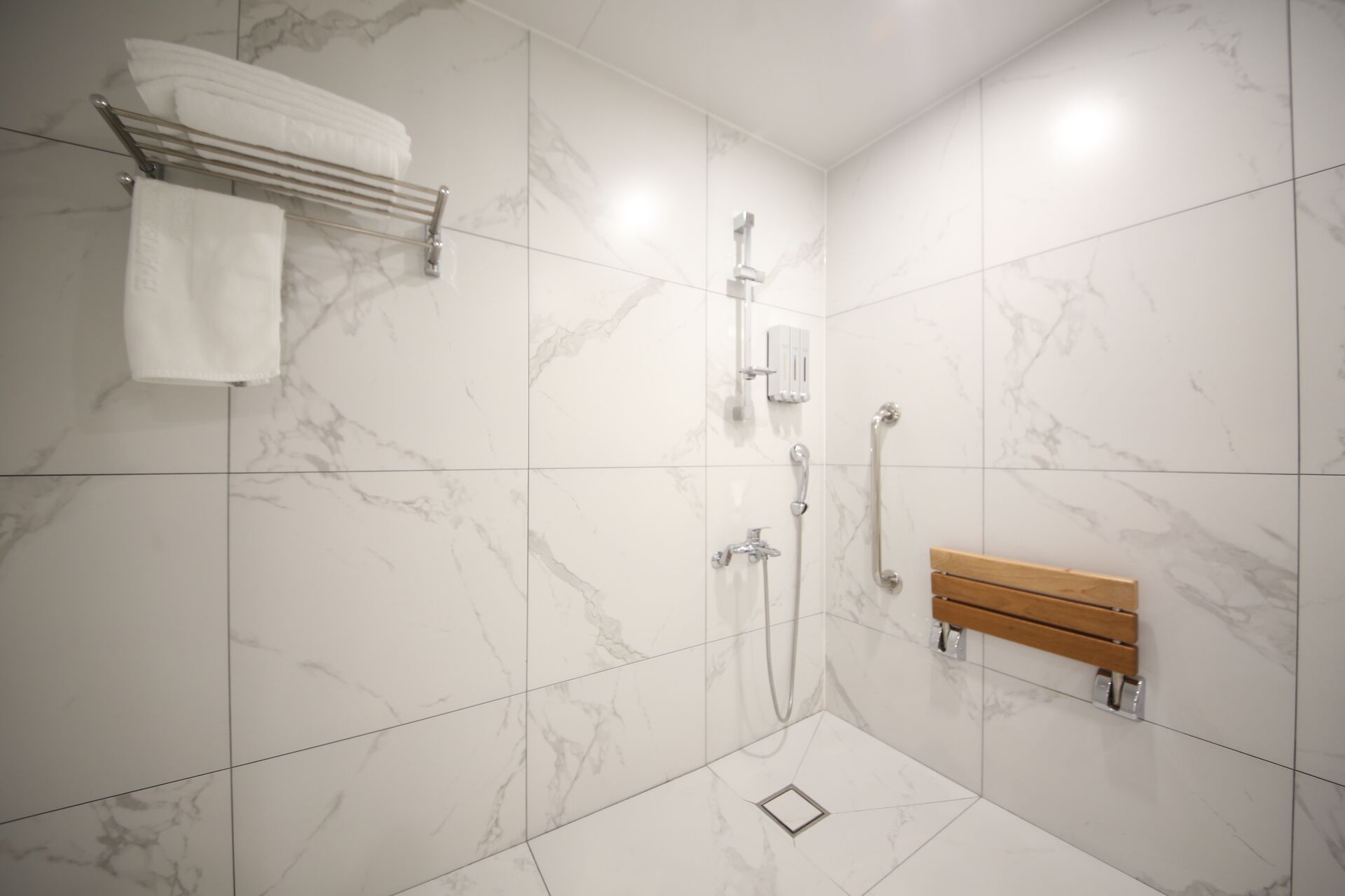 Une salle de bain moderne avec des carreaux de marbre blanc, une douche équipée d’un pommeau de douche à main et d’un banc en bois.