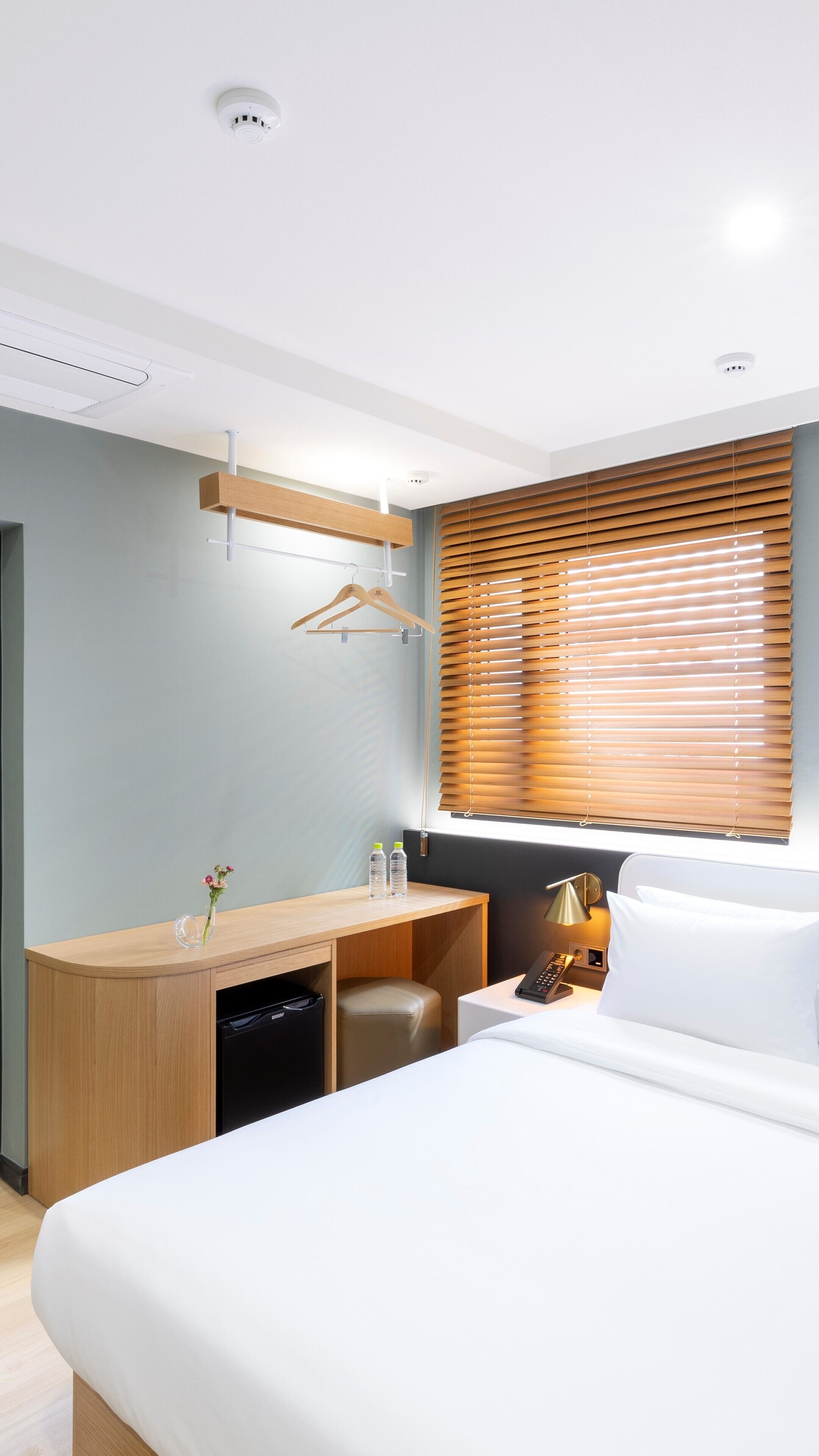 A hotel room with a bed, a desk with a chair, a small table with a lamp, a wooden wardrobe, and a window with blinds.
