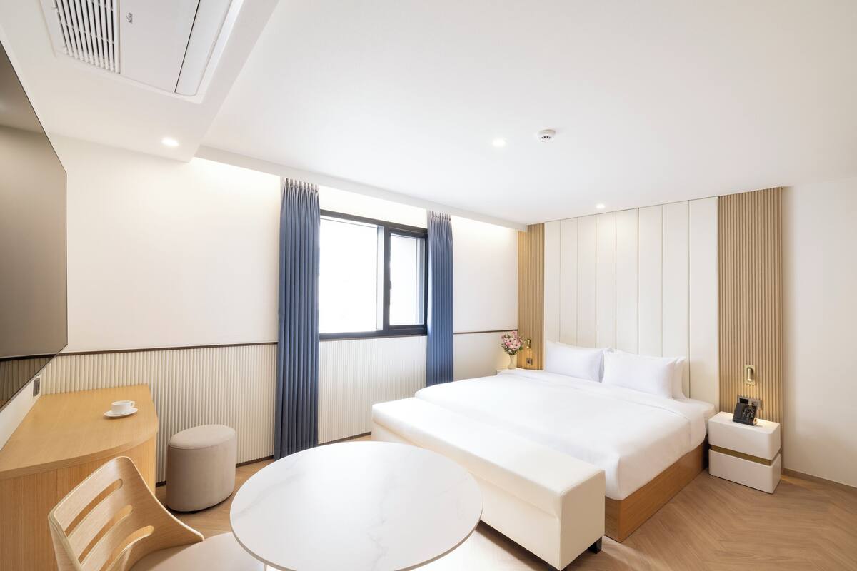 A modern hotel room with a large bed, a round table, a wooden desk, and a small stool.