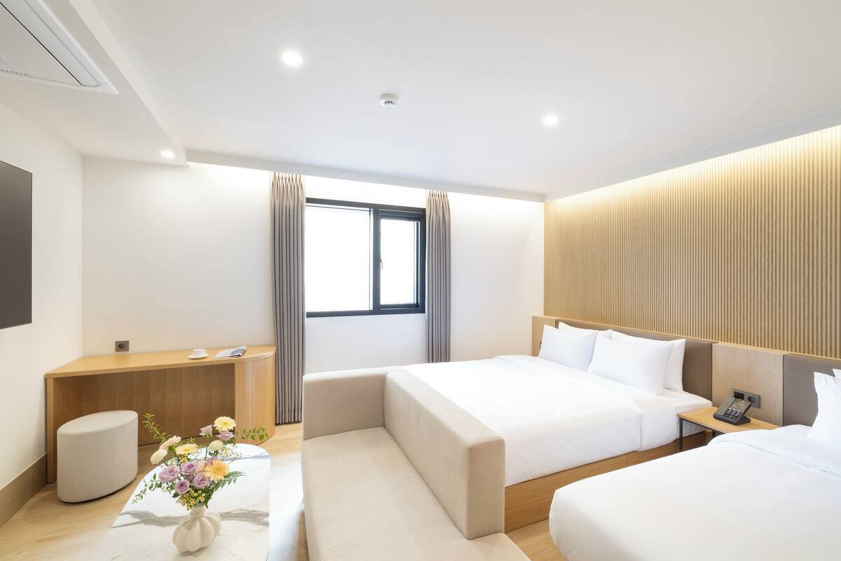 A modern hotel room with a large bed, a bedside table with a phone, a small seating area, a desk, and a window with curtains.