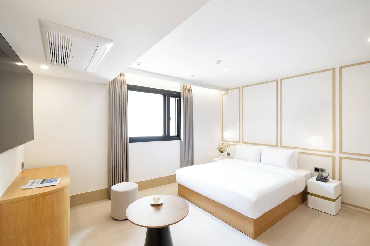 A modern hotel room with a large bed, a bedside table, a small round table, a TV, and a window with curtains.