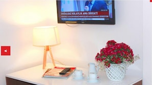 A hotel room with a white table, a lamp, a remote control, a magazine, a vase with red flowers, and a pitcher. - Imerhan Otel (Kusadasi)
