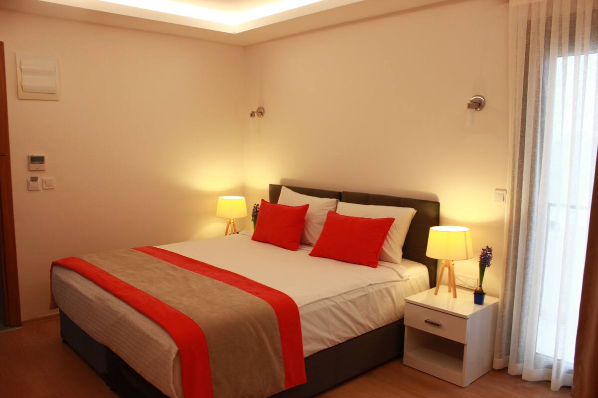 Una cama bien hecha con ropa de cama roja y blanca, dos lámparas de noche y una mesita de noche con lámpara.