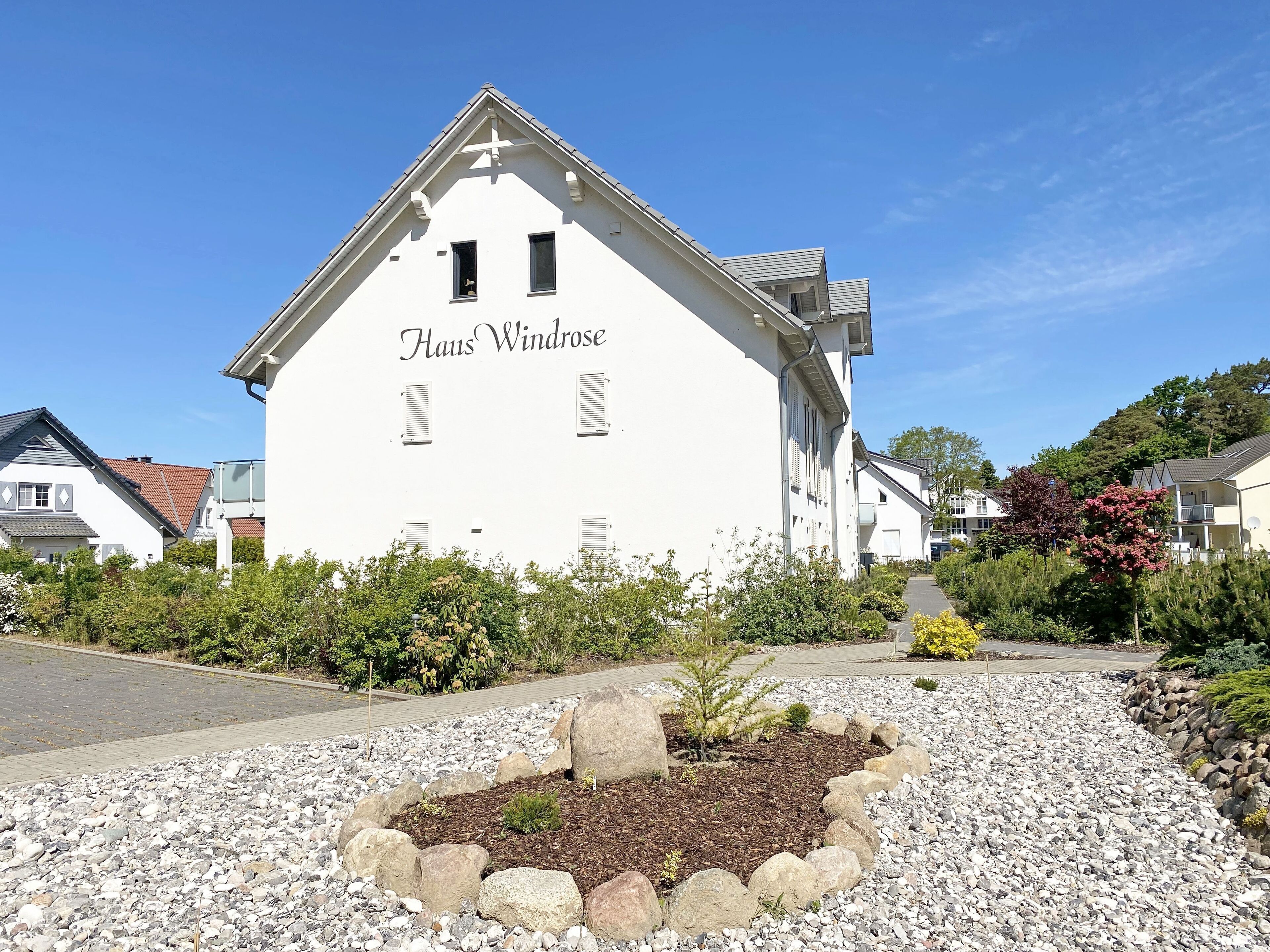 En hvid bygning med navnet "Haus Windrose" på facaden, omgivet af en anlagt have med sten og planter.