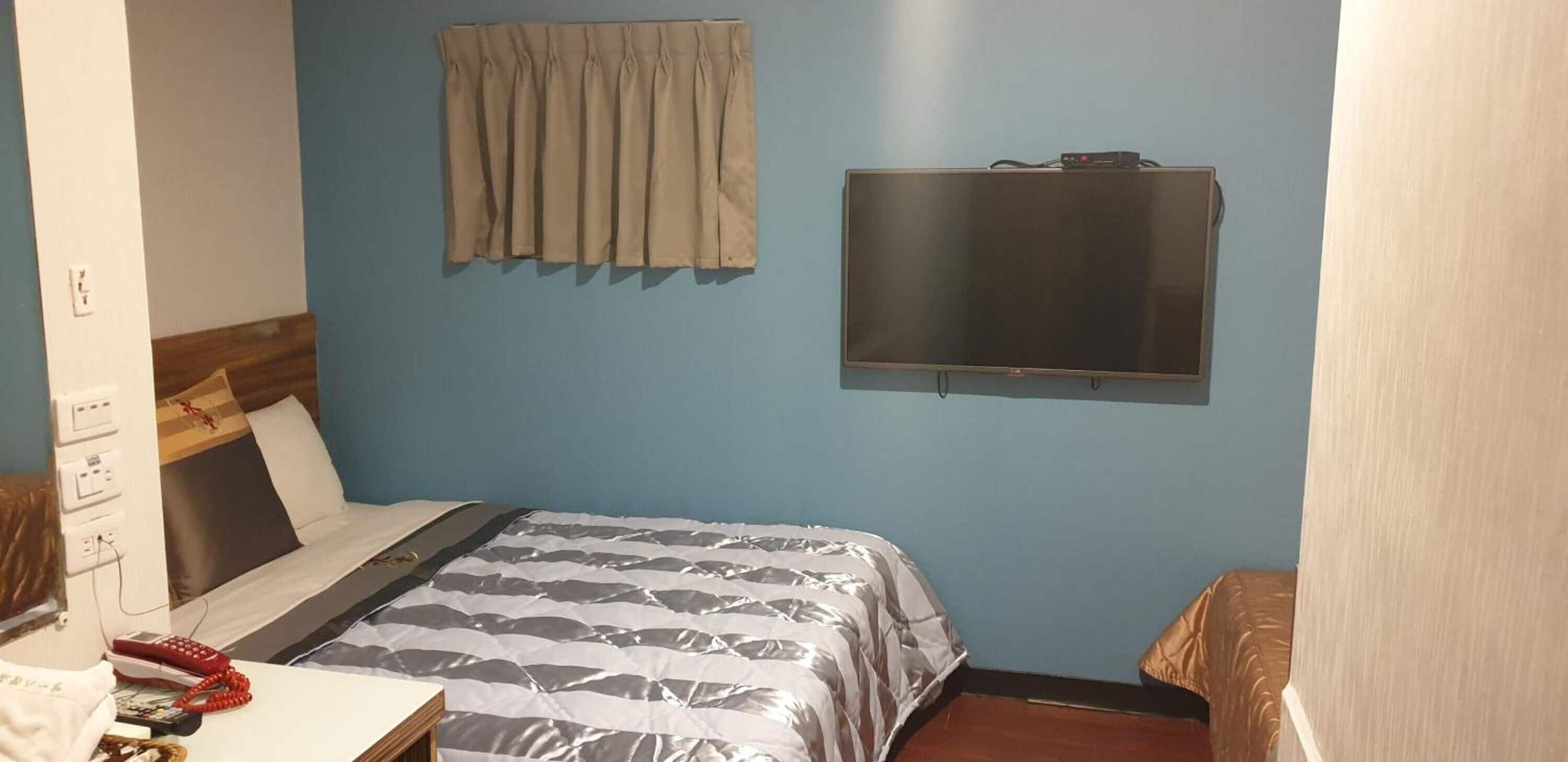 A room with a bed, a television mounted on the wall, a window with curtains, and a bedside table with a telephone and a book.