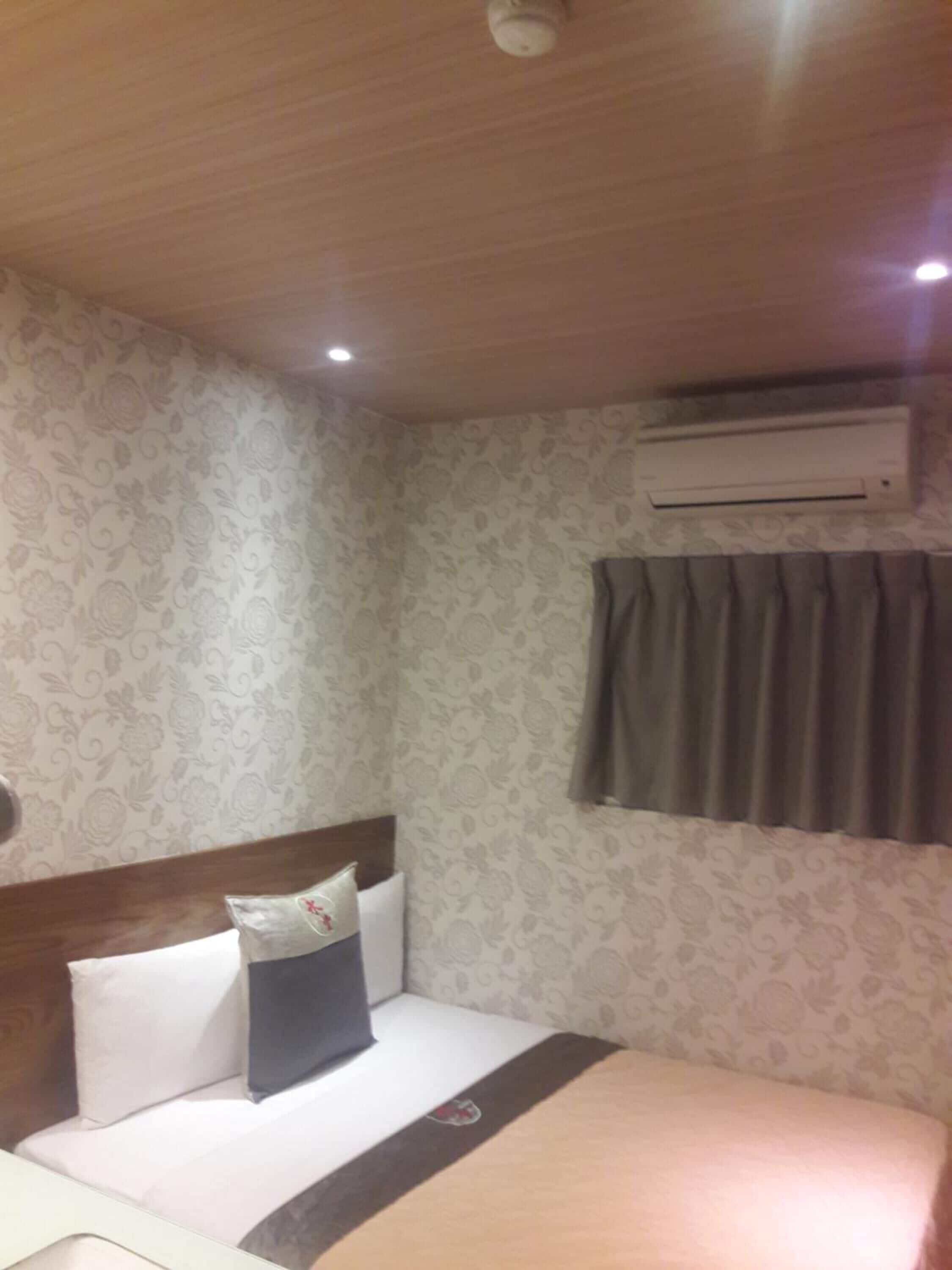 A hotel room with a bed, a headboard, a wall-mounted air conditioner, and a window with curtains.
